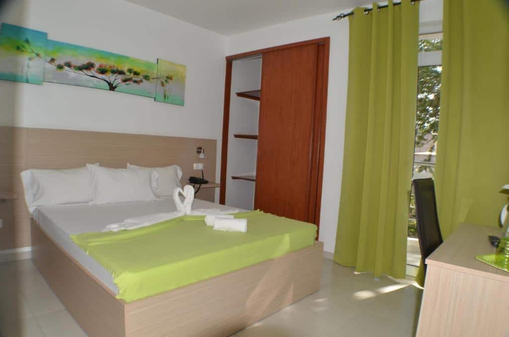 Quarto do Hotel Cesaria em Praia que mostra uma cama de casal e do lado esquerdo da cama um guarda-roupa embutido na parede e do lado uma porta de vidro com uma cortina. No quarto também tem uma escrivaninha e uma cadeira.