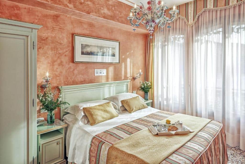 Quarto no Hotel Firenze em tons de rosa salmão com uma parede em mármore, uma janela ampla, um lustre, uma cama de casal com duas mesas de cabeceira com vasos de flores