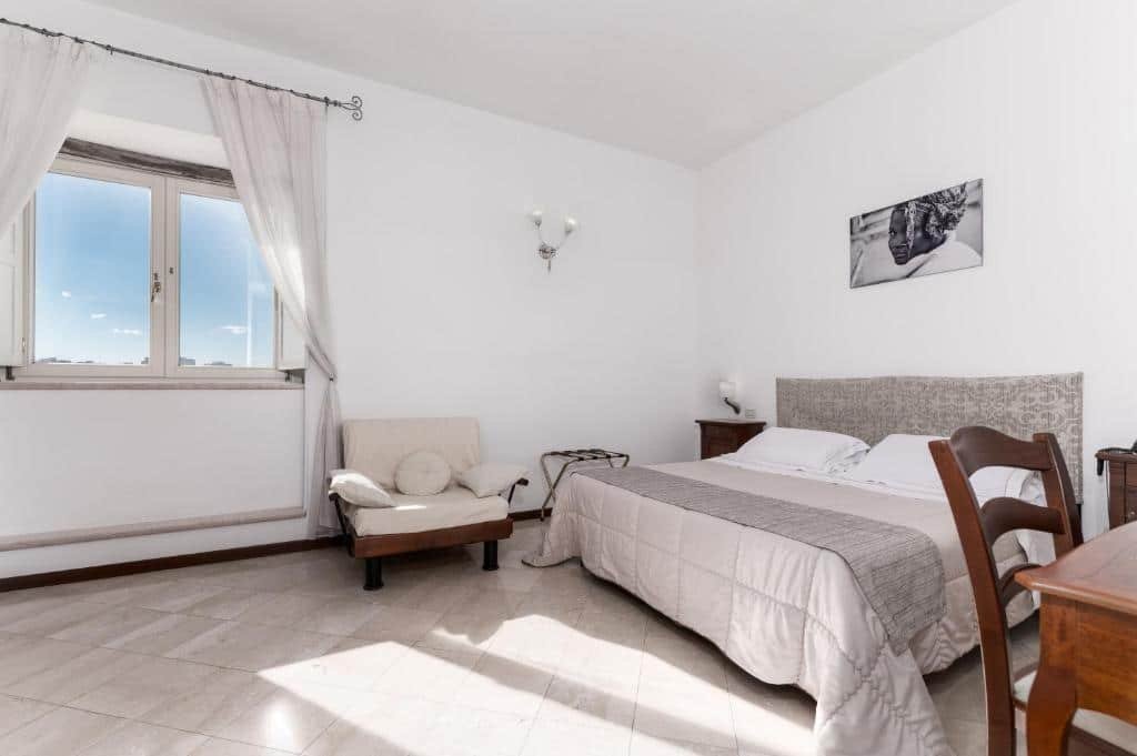 Quarto do Hotel La Terra com uma cama de casal grande com dois travesseiros e um cobertor em cima. Do lado direito tem um pedaço de uma mesa com uma cadeira de madeira e do lado esquerdo tem uma mesinha do lado da cama e uma poltrona com almofadas. Na parede em cima da cama tem um quadro e do lado esquerdo perto da poltrona tem uma janela de vidro fechada com cortinas.