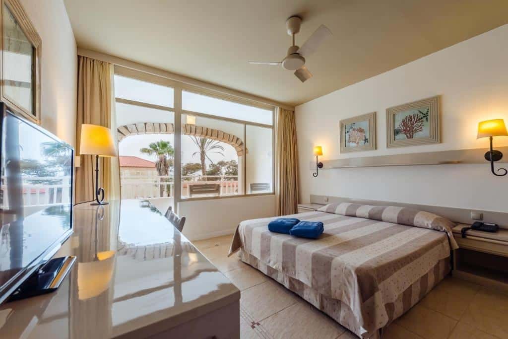 Quarto do Hotel Morabeza em Santa Maria que mostra uma cama de casal e de cada lado da cama uma mesa de cabeceira  e um abajur na parede. Do lado direito tem uma janela grande de vidro com cortinas, um ventilador no teto e em frente uma escrivaninha com uma cadeira, um abajur e uma tv.