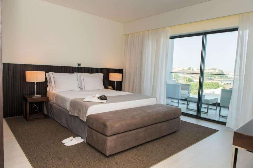 Quarto do Hotel Pérola em Praia que mostra uma cama de casal em cima de um tapete e de cada lado da cama uma mesa de cabeceira com um abajur. Já do lado esquerdo da cama tem uma porta de vidro com cortina que dá acesso a varanda do quarto.