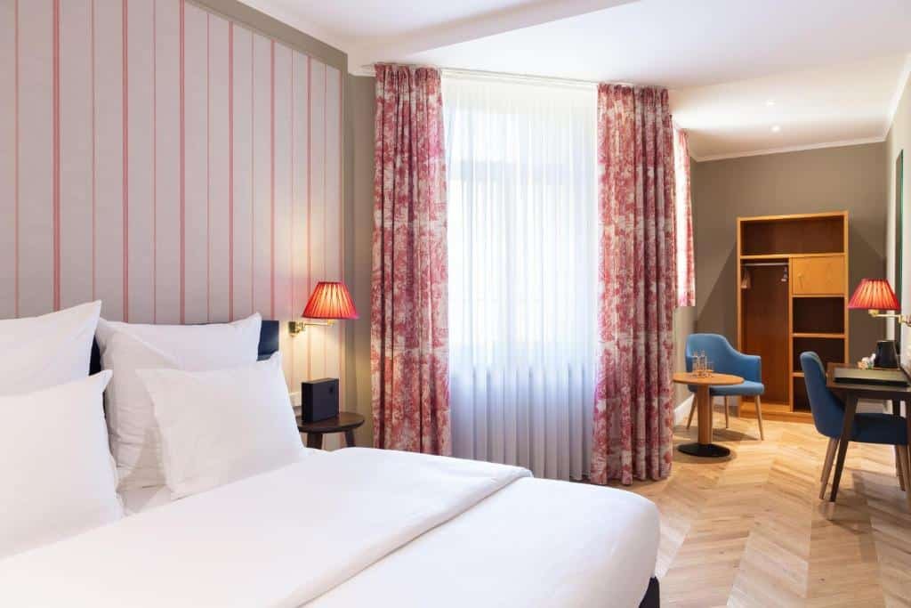 Quarto no Hotel Perrin, para ilustrar o post sobre hotéis em Luxemburgo. A cama é box e de casal, com lençóis brancos, e está posicionada no lado esquerdo da foto. Do seu lado direito, há uma mesa de cabeceira com um abajur vermelho fixo acima. E do lado da mesa de cabeceira, há um janela com cortinas. Já no lado direito da foto, e prosseguindo ao fundo do quarto, vemos alguns móveis de madeira como uma escrivaninha com poltrona, uma meda redonda (também com uma poltrona igual à da escrivaninha), e uma estante de madeira com prateleiras e gavetas atrás, encostada na parede.