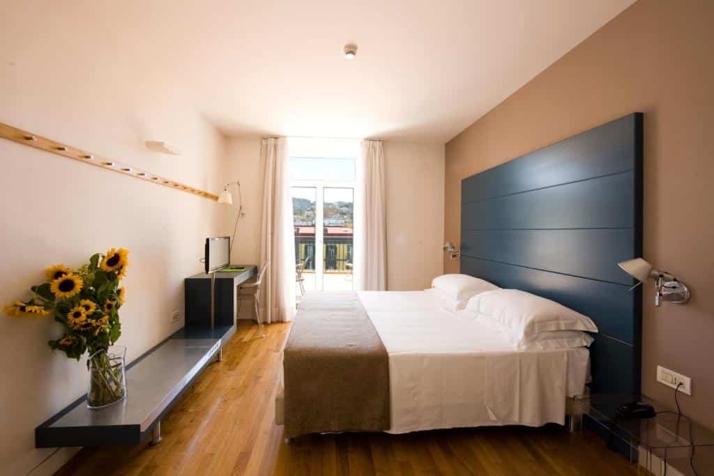 Quarto do Hotel Piazza Bellini & Apartments com uma cama de casal no meio do quarto, dois abajur na parede em cada lado da cama, em frente da cama um banco com um vaso de girassol, ao fundo uma escrivaninha com a tv e ao lado uma porta de vidro com a cortina aberta.