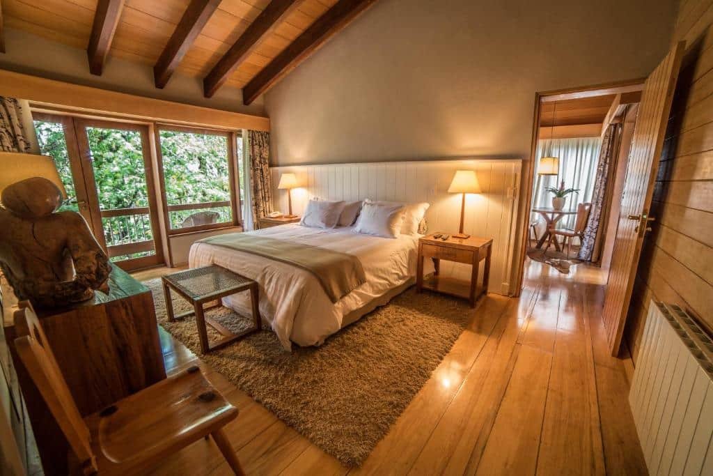 Quarto do Hotel Toriba com chão de madeira, sacada com vista para as montanhas, uma cama de casal, um tapete, duas cadeiras de madeira e duas mesinhas de cabeceira com abajures