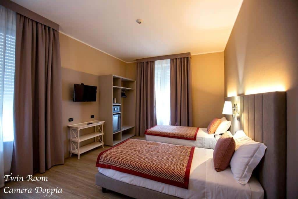 Quarto do Hotel Villa Malpensa com duas camas de solteiro, uma cabeceira com um abajur preso, há um móvel de conceito aberto, uma televisão e duas janelas com cortinas