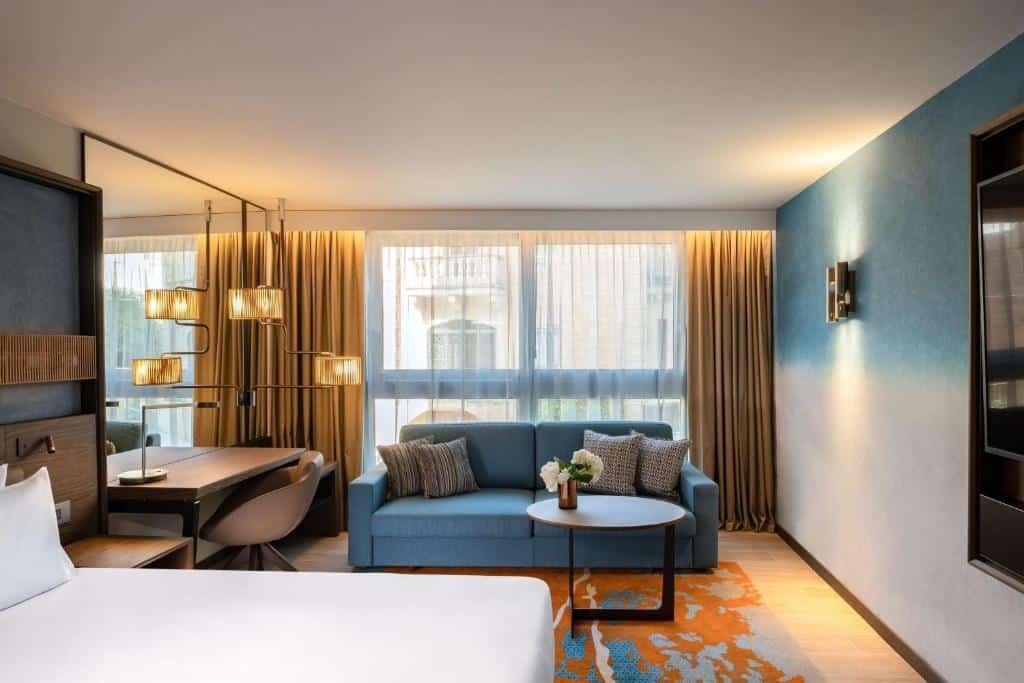 Quarto do Hyatt Regency Malta com uma ampla sacada, um sofá azul com alfmodas, uma cama de casal, um tapete em tons de terra e azul, um espelho, uma mesa de escritório com uma cadeira, e algumas luminárias pelo ambiente