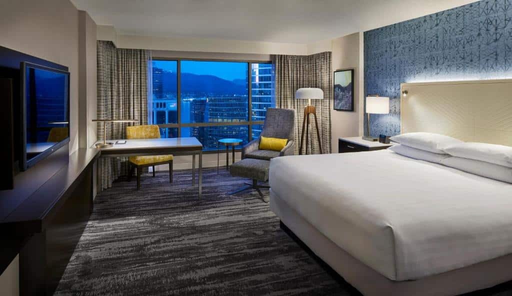 Quarto no Hyatt Regency Vancouver, o local é amplo, há uma janela grande com cortinas e vista para a cidade, uma mesa de escritório com uma cadeira e uma poltrona cinza, o chão é coberto por um carpete cinza, há uma televisão de frente para a cama de casal que tem uma cabeceira branca com iluminação indireta presa nela, a parede atrás da cama tem papel de parede azul com detalhes preto