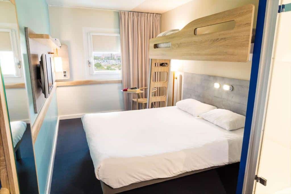 Quarto do ibis Budget Luxembourg Aeroport, para ilustrar o post sobre hotéis em Luxemburgo. A cama é box de casal, e acima dela, há uma beliche com uma cama de solteiro. Do lado da escada da beliche, há uma cadeira, e atrás dessa cadeira, está a janela com uma cortina bege. Na frente da cama de casal, há uma TV fixa na parede e um espelho ao seu lado.