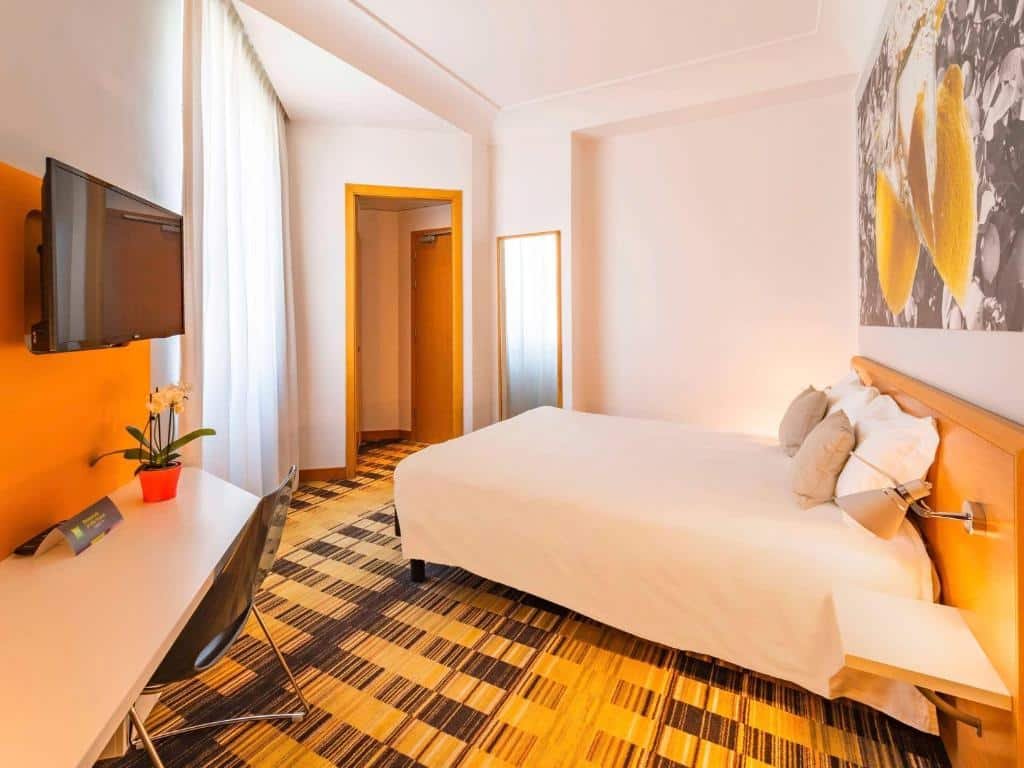 Quarto do Ibis Styles Napoli Garibaldi com uma cama de casal em baixo de um quadro grande parede, ao lado da cama uma mesinha com abajur, em frente uma escrivaninha com uma cadeira e uma tv e ao fundo um espelho e uma porta de madeira.