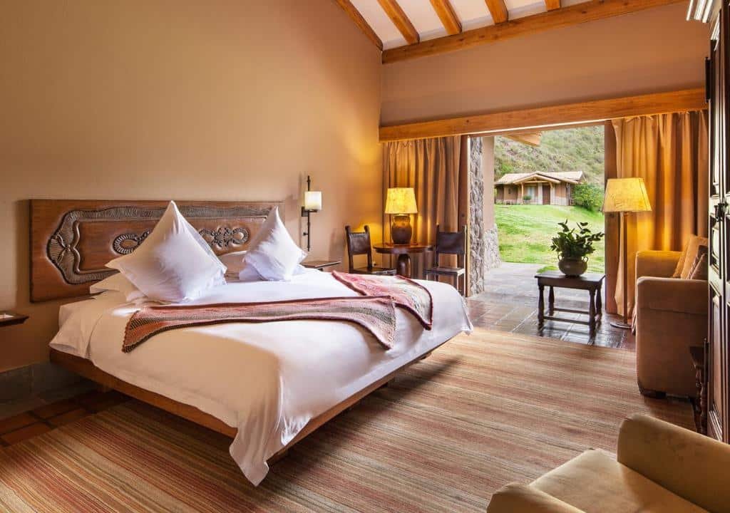 suíte do Inkaterra Hacienda Urubamba com uma ampla cama de casal no lado esquerdo do quarto, ao lado de uma pequena mesa de madeira redonda. Ao fundo há uma varanda terrestre com acesso ao jardim do hotel.