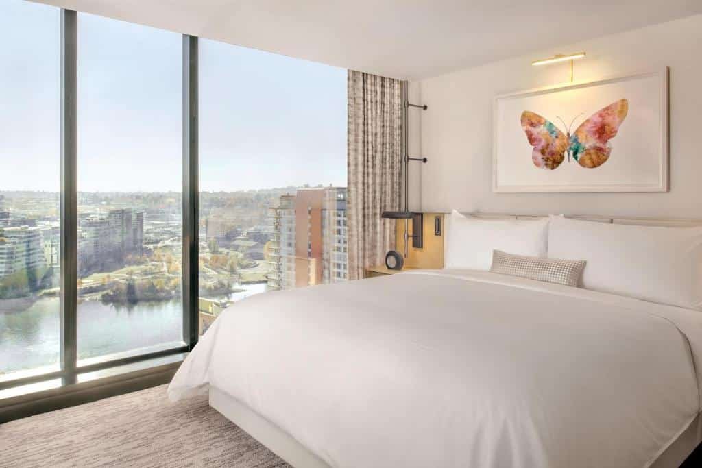 Quarto no JW Marriott Parq Vancouver com uma ampla janela que vai do texto até o chão dando vista para uma baía e outro prédios, dentro do quarto há uma cama de casal e sob ela há um quadro com uma borboleta desenhada, as paredes são brancas e o piso tem carpete em tom de bege