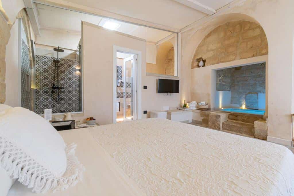 Quarto da Masseria dei Monaci com uma cama de casal com travesseiros, em frente uma parede de pedras com uma escada de pedras também que dá acesso a banheira do quarto. Já ao lado esquerdo da cama tem um vidro que mostra o chuveiro, do lado tem uma porta de vidro que dá acesso a outros cômodos do quarto e ao lado uma parede com uma tv e uma mesinha em baixo. 