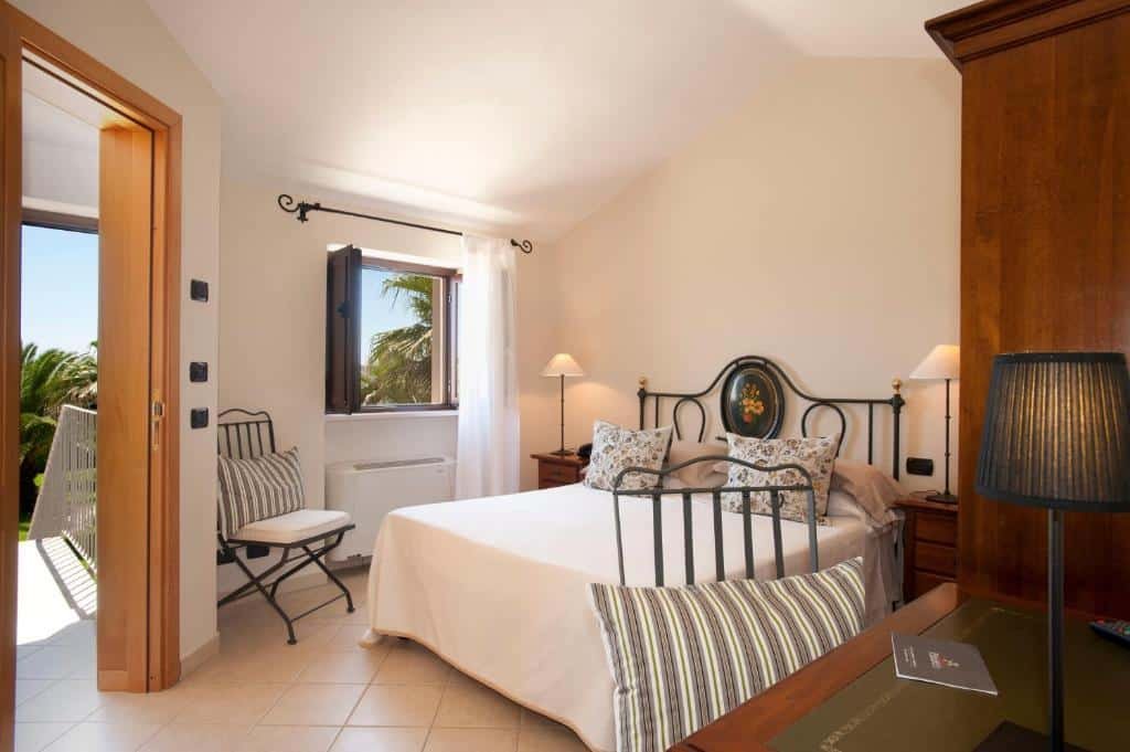 Quarto da Masseria Panareo com uma cama de casal com travesseiros e almofadas em cima, do lado direito tem um móvel de madeira e parte de uma escrivaninha com um abajur e uma cadeira aparecendo. Já na parte esquerda do quarto tem uma cadeira, uma janela aberta com vista para o céu azul e as palmeiras, e tem também uma porta de madeira aberta com vista para a paisagem.