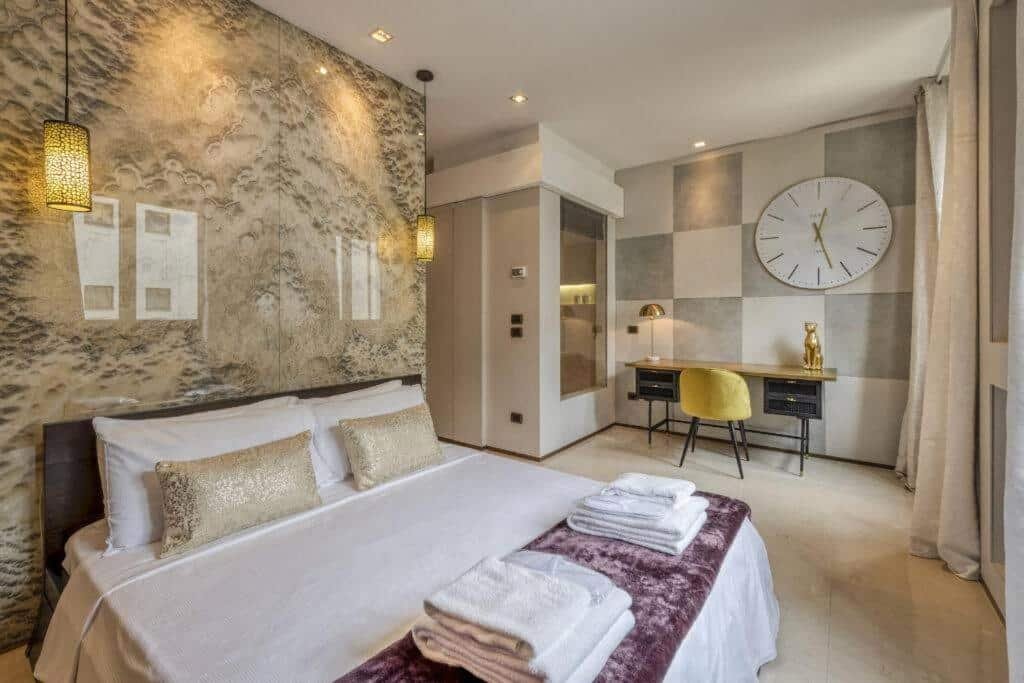 Quarto do Milan Royal Suites - Centro com uma cama de casal com almofadas e toalhas, há duas luminárias presas no teto, há também uma mesa de escritório com uma cadeira, além de uma sacada com cortinas, para representar onde ficar em Milão