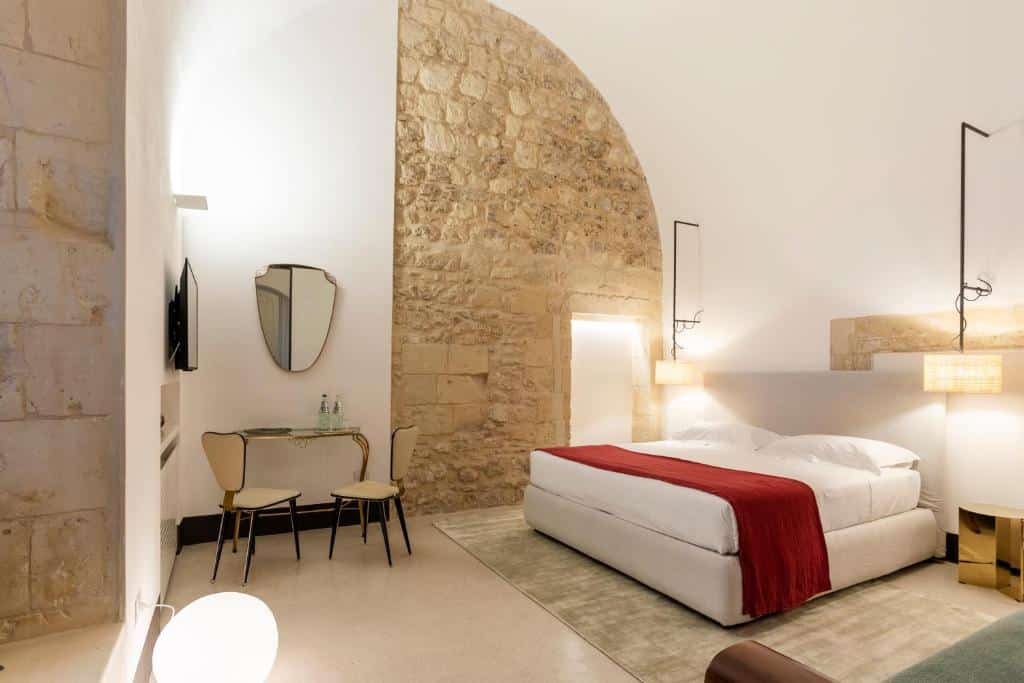 Quarto do Palazzo De Noha - Boutique Hotel com uma cama de casal ao centro com dois lustres de cada lado da cama. Já a parede ao fundo do quarto é metade com pedras a vista e a outra parte é pintada de branco com um espelho e uma tv nela e uma mesa com duas cadeiras perto.