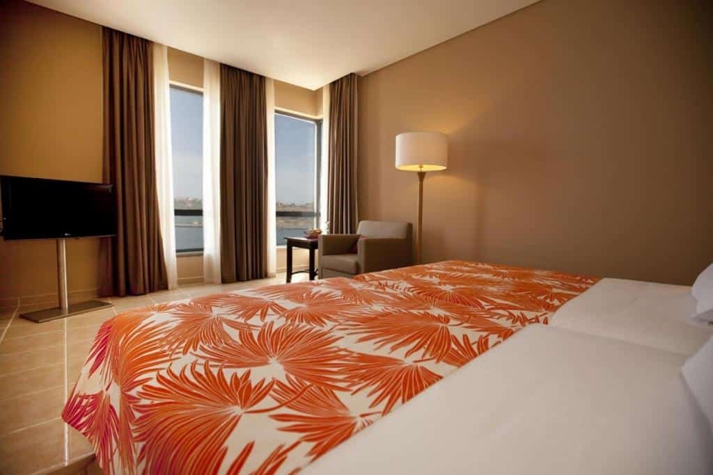 Quarto do Pestana Tropico Ocean & City Hotel em Praia que mostra uma cama de casal e em frente a cama tem uma tv, duas janelas de vidro com cortinas, uma poltrona com uma mesa e um abajur de chão.