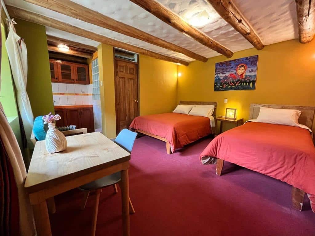 quarto do Pisac Inca Guest House com duas camas de casal, dispostas lado a lado, com uma mesa de amdeira para trabalho em frente as camas. Há ainda um espaço com cozinha compacta dentro do quarto.