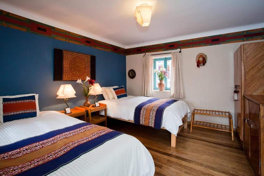 quarto do Pisac Inn com duas camas de solteiro, dispostas lado a lado, no canto esquerdo da imagem. Há duas escrivaninhas entre as camas, com abajures em cada uma.