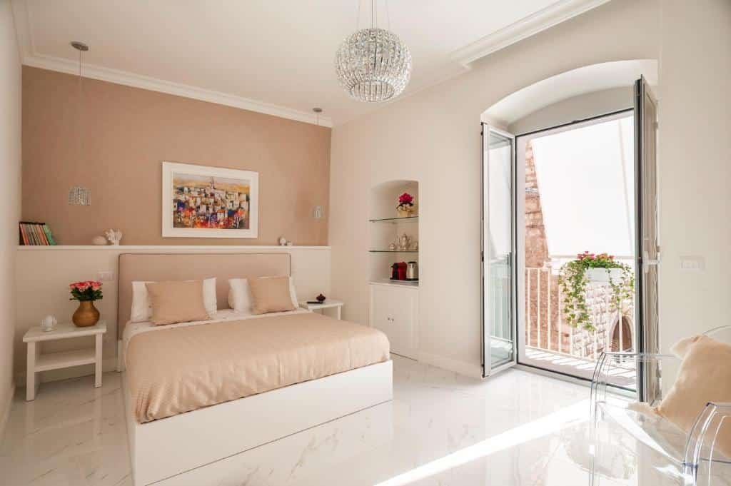 Quarto do Porta Piccola Luxury Home com uma cama de casal ao centro, do lado esquerdo tem uma mesinha com um vaso de flor e do lado direito tem algumas prateleiras com objetos. Na parede atrás da cama tem um quadros, dois lustres e alguns livros, já em frente a cama tem uma poltrona transparente  com uma almofada e uma porta de vidro que dá acesso a varanda do quarto.