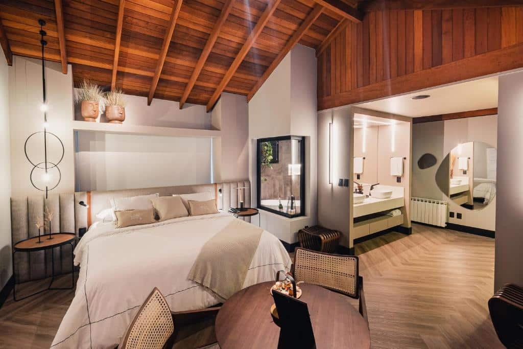 suíte espaçosa da Pousada Bella Terra em Gramado com uma cama de casal à esquerda, uma mesa redonda de madeira com duas cadeiras à frente da cama, e no lado direito, duas pias brancas com acesso ao banheiro que possui uma janela de vidro, de onde é possível ver a banheira