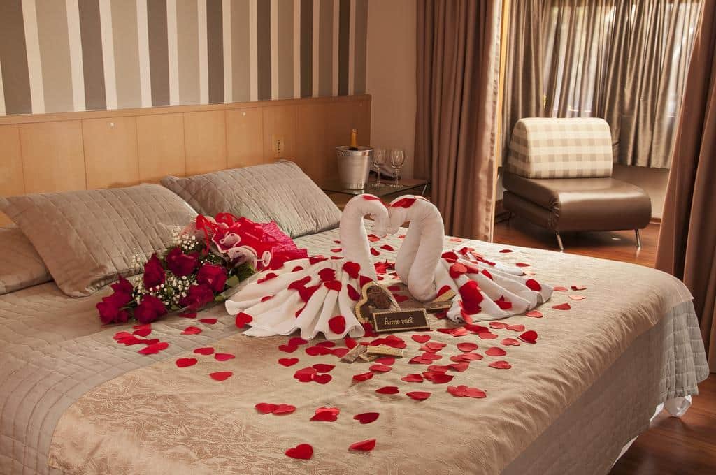 cama de casal da Pousada Bernardete com pétalas de rosa espalhadas sobre a cama, além de um buquê deitado, juntamente com dois origamis de toalhas brancas em formato de cisnes de frente um para o outro.