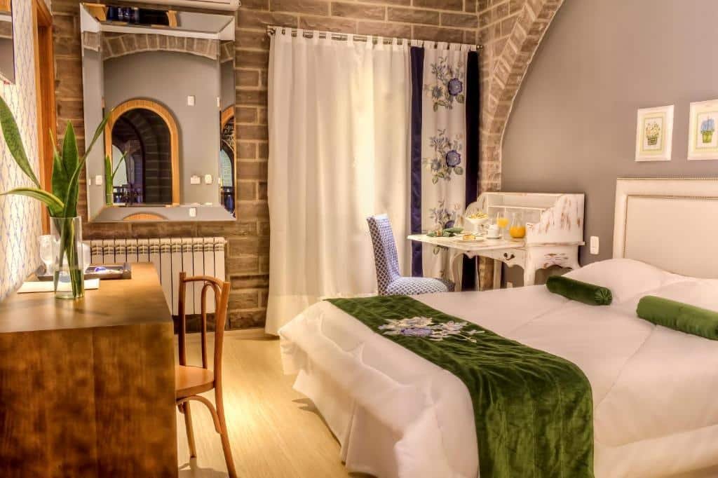 quarto do Pousada Castello Benvenutti com cama de casal, penteadeira expansiva com uma refeição servida sobre ela, mesinha de trabalho, espelho sobre o aquecedor, e cortina