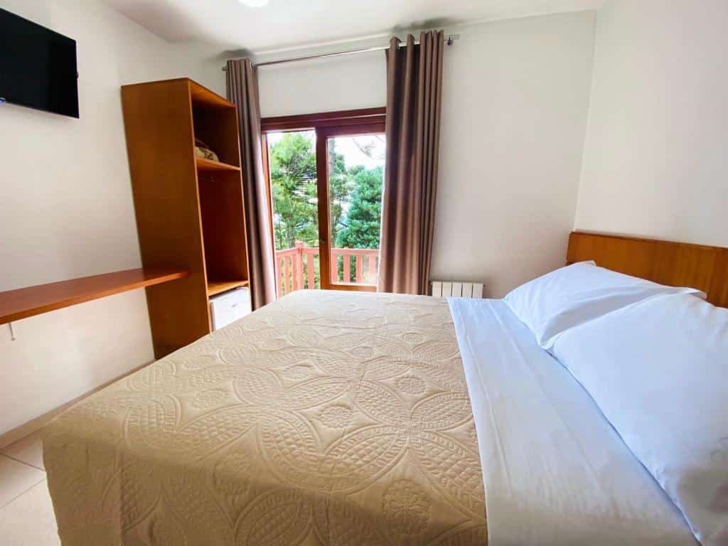 quarto da Pousada do Lago Gramado com uma cama de casal no lado direito e uma porta para a sacada no canto esquerdo. A visão lá de fora é de várias araucárias