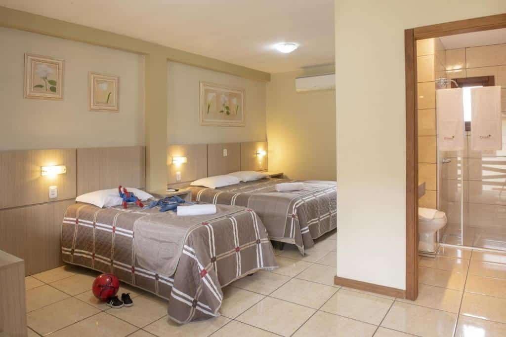 suíte da Pousada Floratta Nossa Casa em Gramado com uma cama de casal e uma cama de solteiro, onde um boneco do homem aranha está jogado em cima do travesseiro, e no chão há uma bola vermelha com dois tênis pretos pequenos. No lado direito da imagem há uma porta aberta que dá acesso ao banheiro com box.