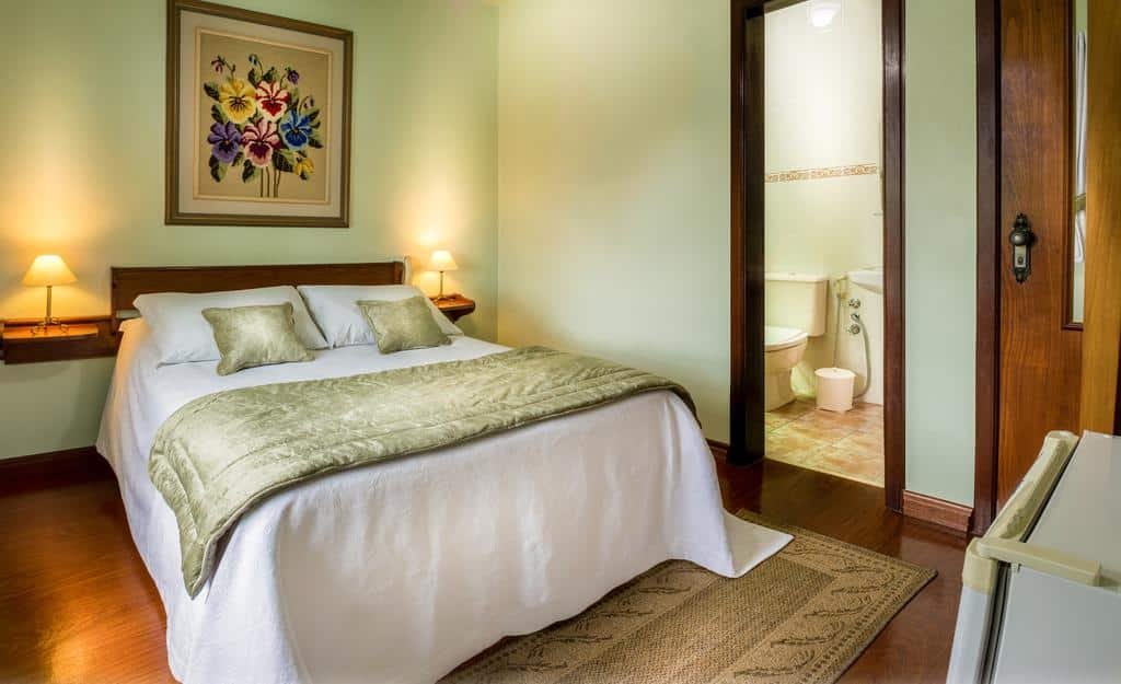 suíte da Pousada Mikolay com uma cama de casal à esquerda e uma porta com acesso ao banheiro no lado direito. Sobre a cama há uma quadro de flores coloridas