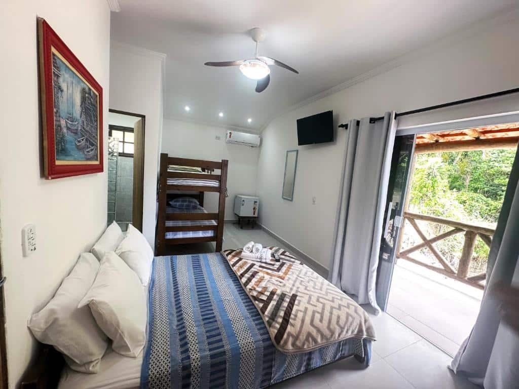 Quarto da Pousada Sobre As Ondas de Picinguaba com uma cama de casal de frente para uma porta de vidro que dá acesso a varanda do quarto. Ao lado esquerdo da cama ao fundo do quarto tem um beliche de madeira, uma porta que dá acesso ao banheiro e do lado do beliche um ar-condicionado, um frigobar, um espelho e uma tv.