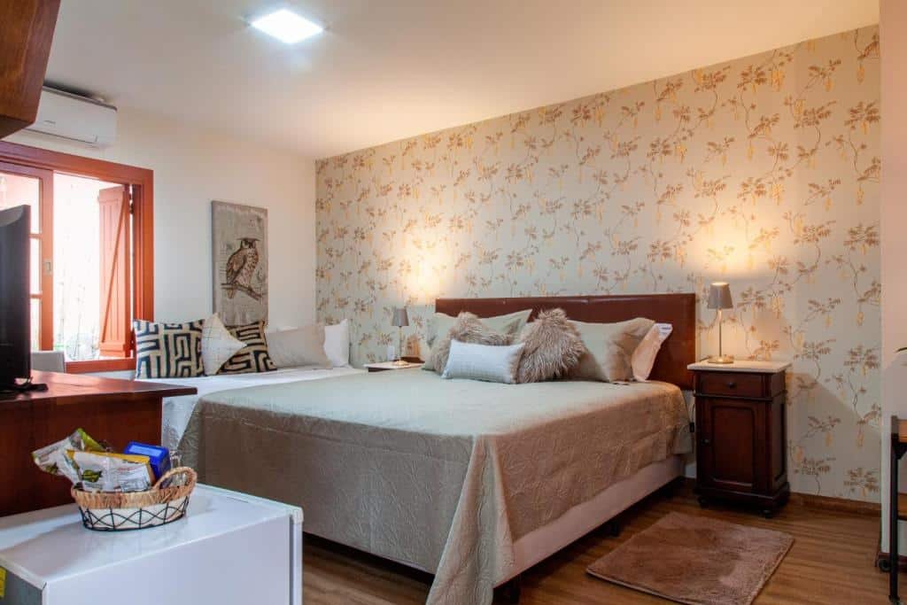 Quarto da Pousada Vila 505 com cama de casal, duas cômodas de madeira com luminária, um sofá cama do lado esquerdo, TV em frente a cama em cima de uma cômoda e frigobar do lado da TV.