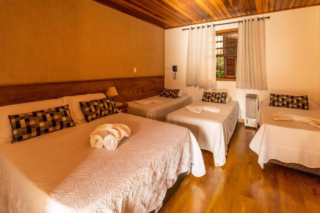 Quarto da Pousada Vila Floratta com três camas de solteiro, uma cama de casal, uma janela com cortinas e um ar-condicionado de chão por entre as camas, para representar onde ficar em Campos do Jordão