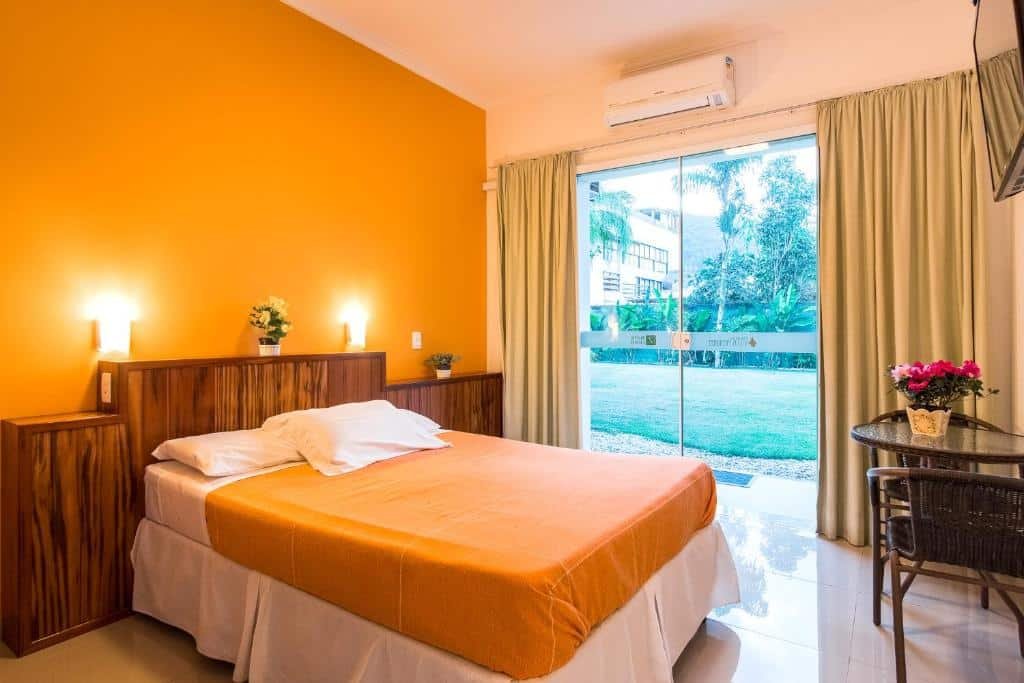 Quarto na Pousada Villa Piemonte na Praia das Toninhas, uma quarto com uma cama de casal, tudo em tons de amarelo, iluminação indireta sob a cama, alguns pequenos vasos de flor, uma televisão, uma mesa com duas cadeiras, uma porta de vidro virada para o jardim com cortinas, e um ar-condicionado, para representar pousadas na Praia das Toninhas