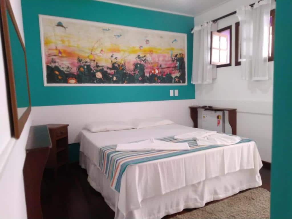 Quarto do Pousada Vison com uma cama de casal, duas janelas com cortinas, um tapete, um espelho e dois móvels de madeira, além de um frigobar