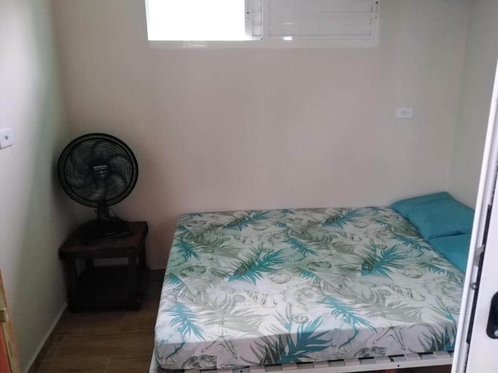 Quarto do Recanto do Estaleiro com uma cama de casal com dois travesseiros e um lençol, uma janela aberta na parede e um ventilador em cima de uma mesinha.