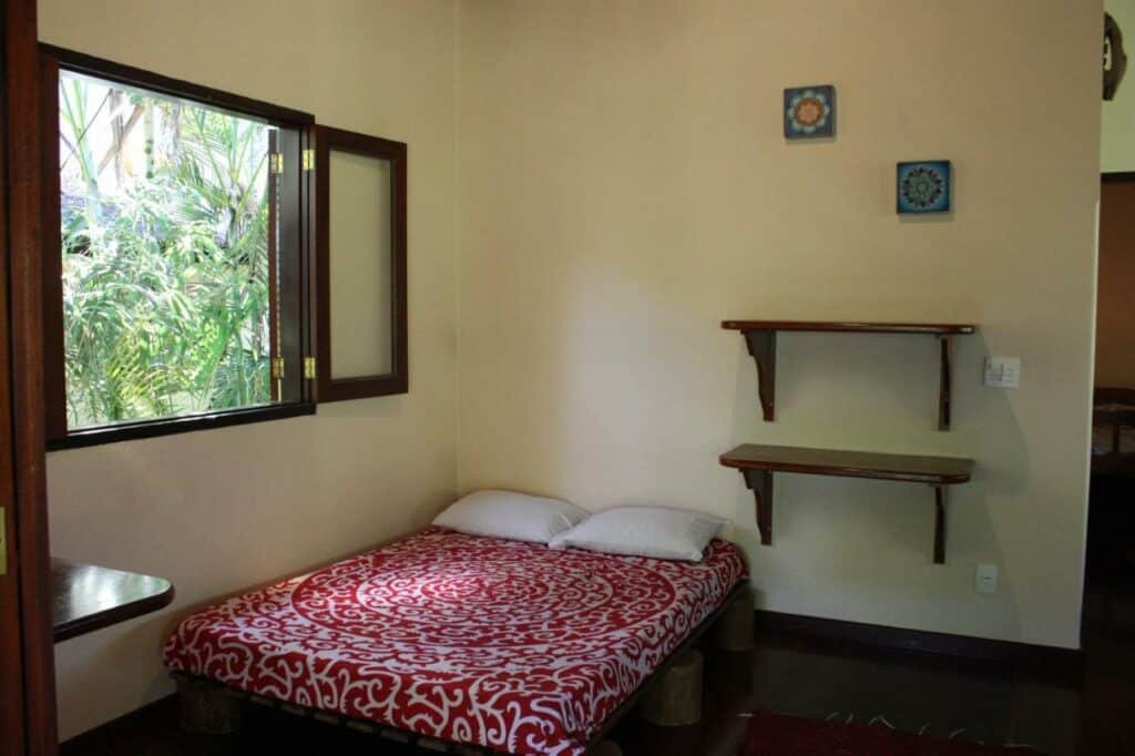 Quarto do Recanto Tangara – Casa – Praia do Felix com cama de casal do lado esquerdo da imagem no canto debaixo de uma janela ampla de madeira e do lado direito da cama em cima duas prateleiras de madeira. Representa airbnb na praia do Félix.