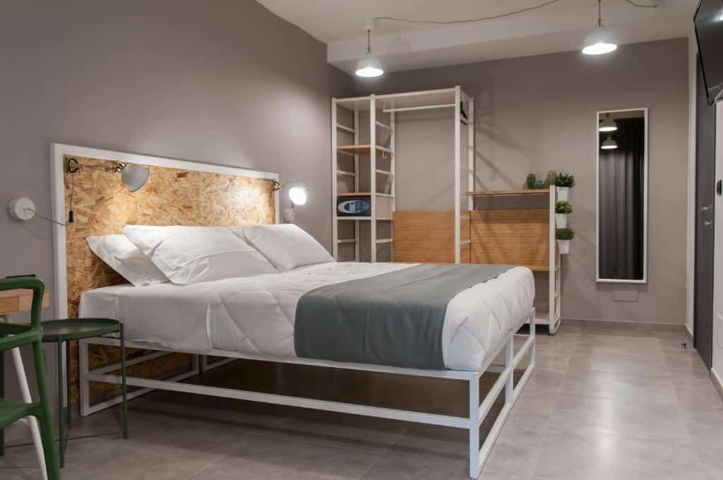 Quarto do Residence Hotel Moderno com uma cama de casal com a cabeceira de madeira, dois abajures de parede em cada lado da cama e do lado esquerdo tem uma mesinha verde redonda e parte de uma cadeira. Já do lado direito da cama tem um guarda-roupa aberto, com um cofre, algumas plantas pequenas e do lado um espelho na parede.