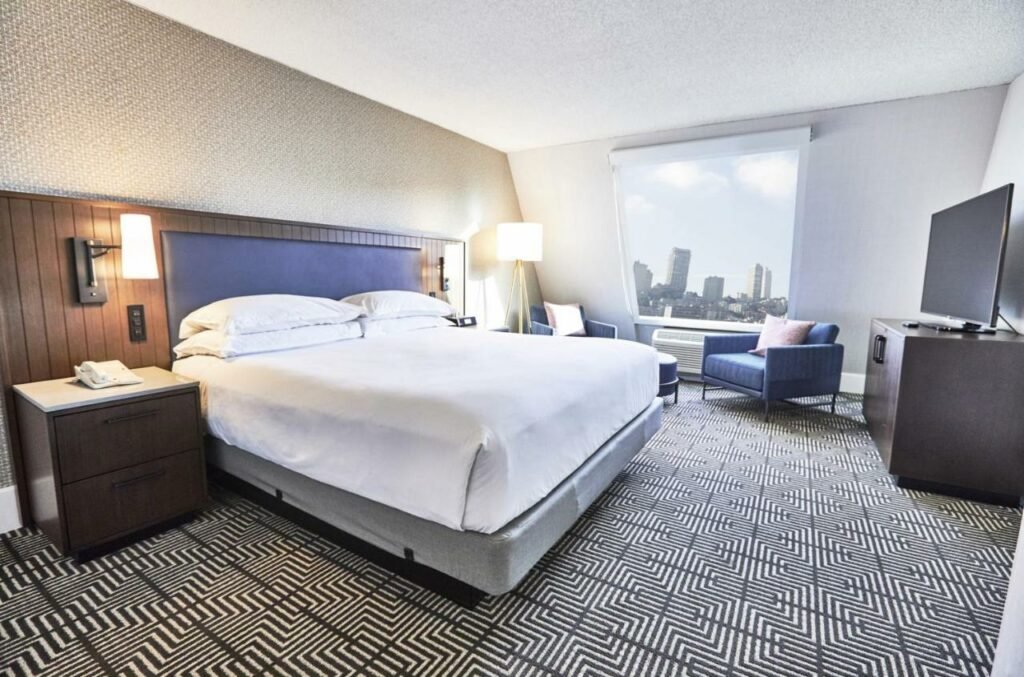 Quarto do Riu Plaza Fisherman’s Wharf com cama de casal do lado esquerdo da imagem e uma cômoda em cada lado da cama. Do lado esquerdo da cama há uma luminária grande, perto de duas poltronas que ficam logo abaixo da janela. Uma TV está perto delas e de frente para a cama. Representa hotéis em San Francisco.