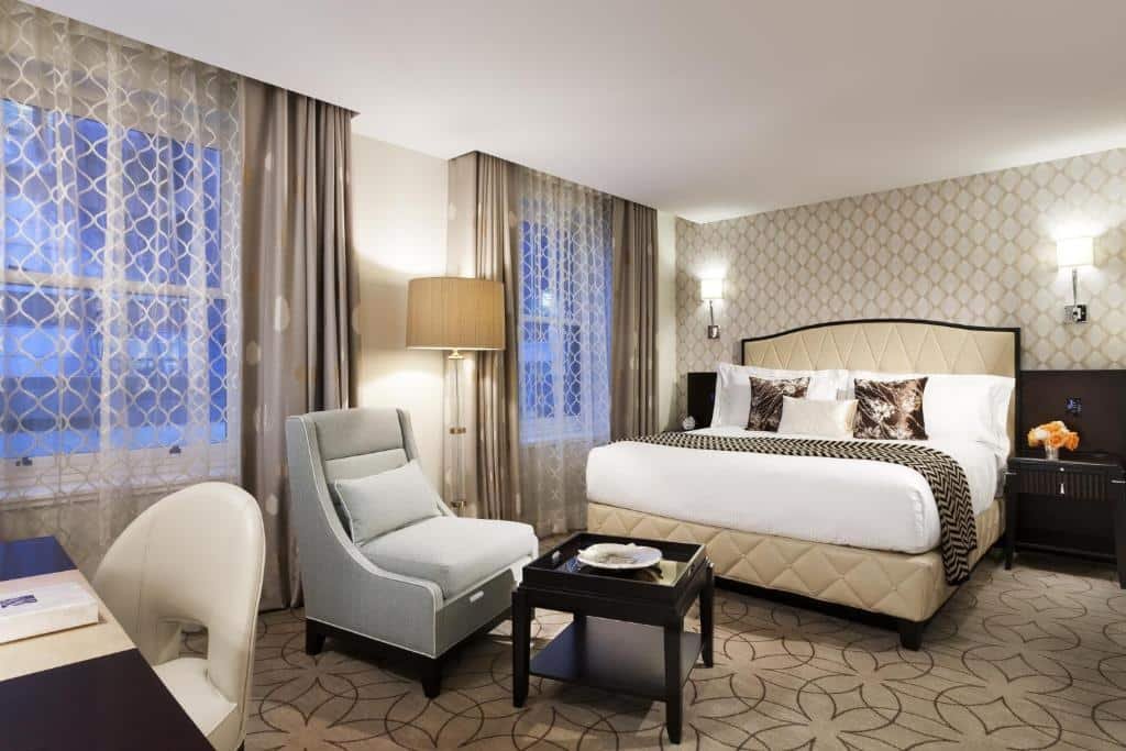 Quarto do Rosewood Hotel Georgia, há duas janelas com cortinas, uma cama de casal com almofadas, na frente da cama tem uma poltrona cinza, o chão é de carpete bege com desenhos minimalistas em marrom, o quarto é inteiro decorado em tons de bege e tons de madeira