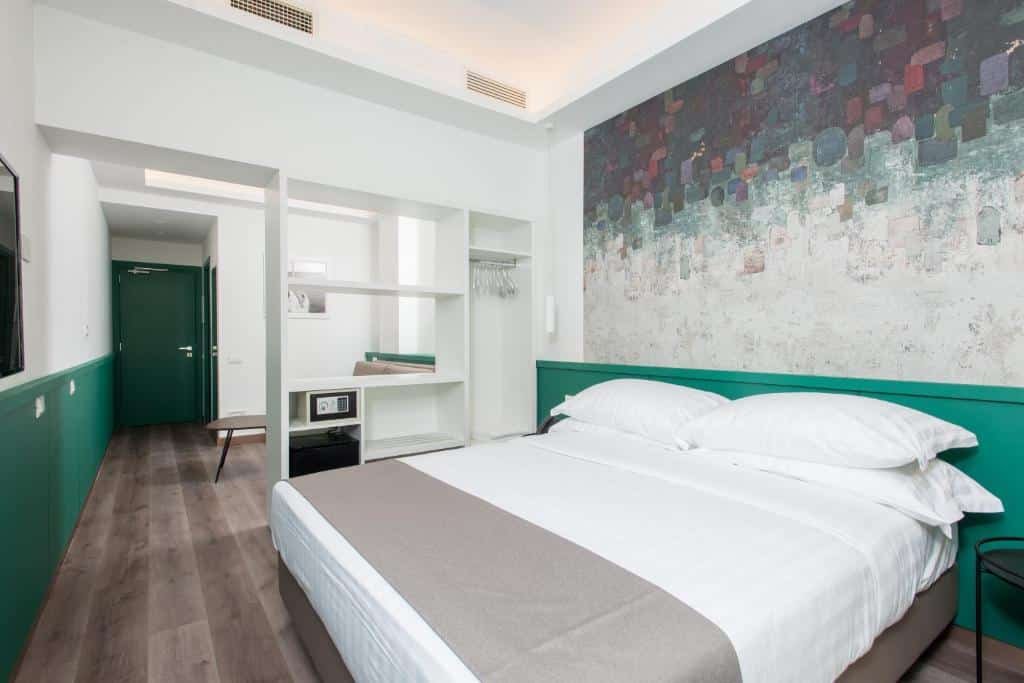 Quarto do Smart Hotel Napoli com a cama bem no foco da imagem e ao fundo um armário aberto com cabides, um pequeno cofre e espaços vazios para colocar seus objetos. Mais ao fundo uma porta verde de entrada.