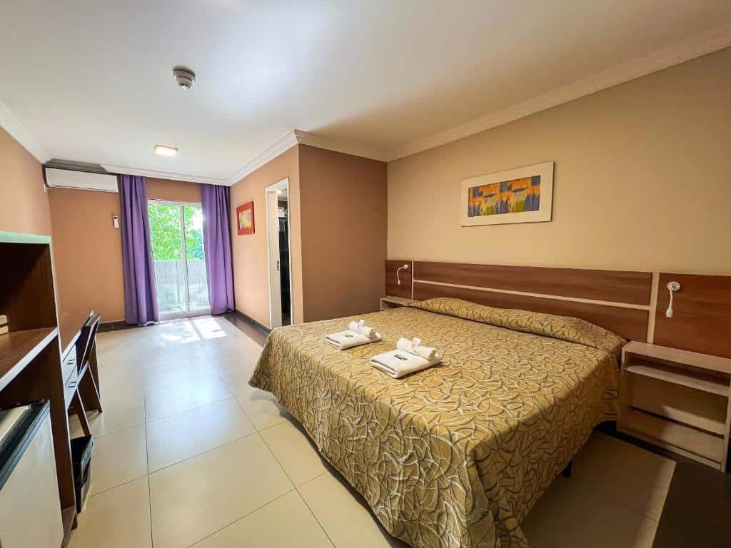 Quarto do Suíça Hotel & Resort com uma cama de casal do lado direito do quarto e na frente da cama tem uma armário de madeira e uma mesa com cadeira. Ao fundo do quarto do quarto tem uma porta que dá acesso ao banheiro e mais ao fundo uma porta de vidro com cortina que dá acesso a varanda.