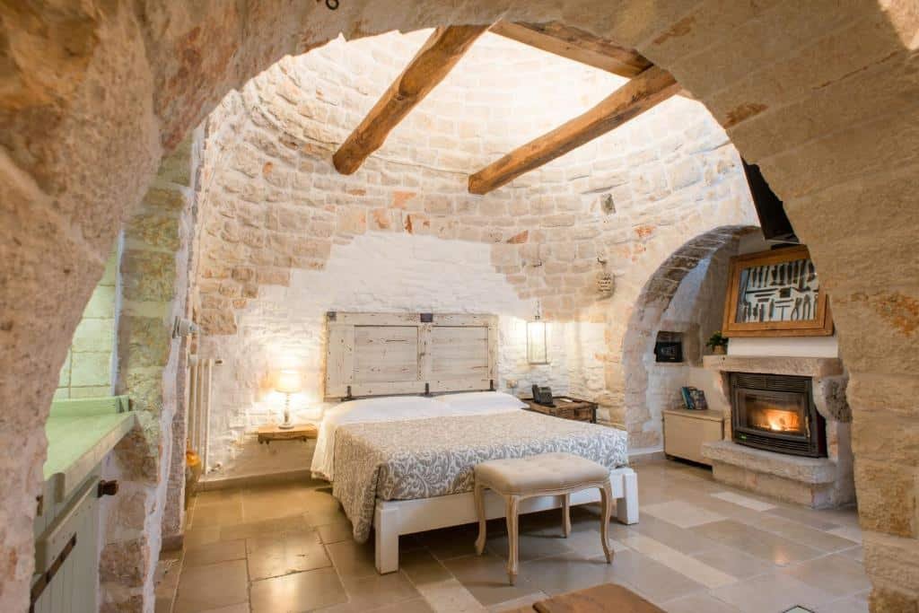 Quarto do Trulli Holiday Albergo Diffuso com uma cama de casal ao centro com um banco estofado no pé da cama, e uma mesinha de cada lado da cama, uma com um abajur e a outra com um telefone. Do lado direito da cama tem uma fogueira fechada com alguns móveis do lado e um quadro em cima. O quarto é feito de paredes de pedra e tem um formato parecendo uma caverna.