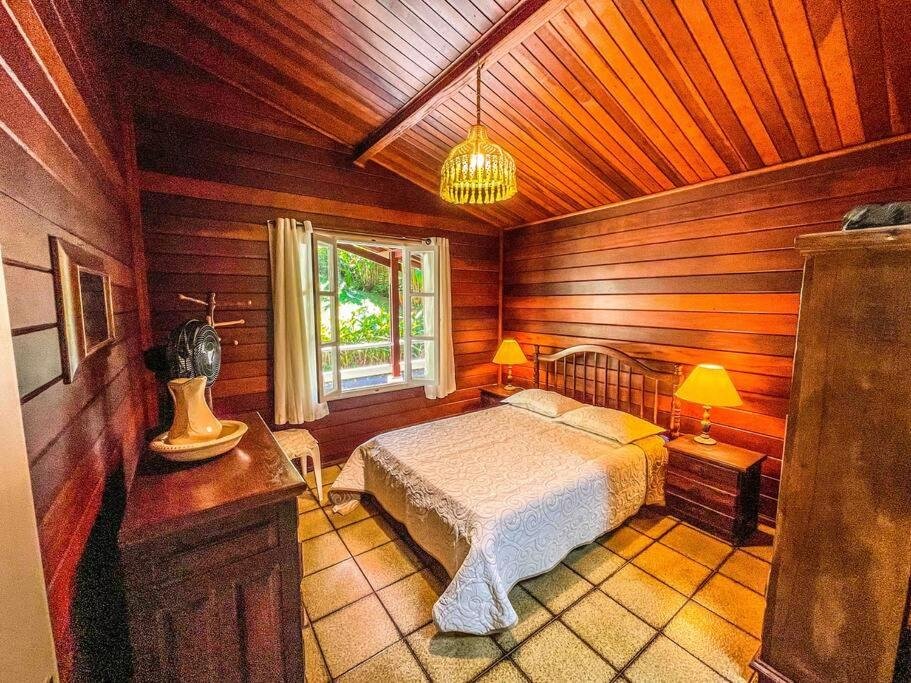 Quarto do Ubatuba, SP- Brasil- Praia do Felix – Casa do Aconchego com cama de casal do lado direito da imagem no centro do quarto com uma cômoda em casa lado com luminária em frente a cama uma cômoda de madeira.