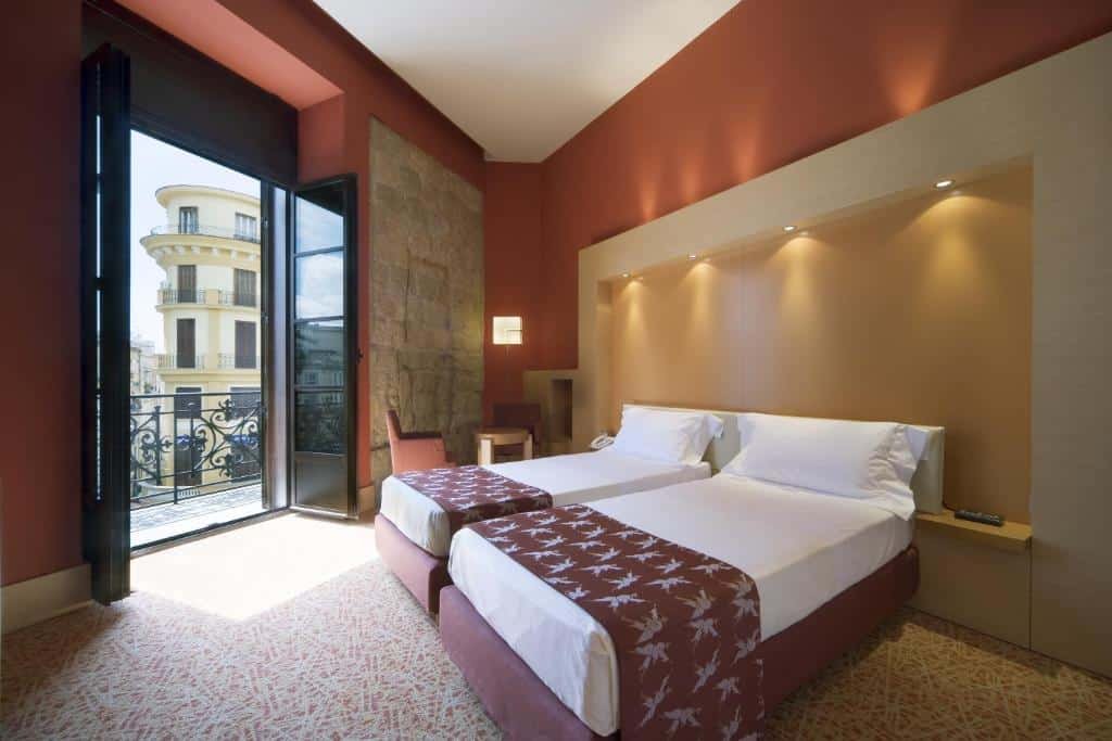 Quarto do UNAHOTELS Napoli com duas camas de solteiro bem próximas no centro do quarto, já no canto esquerdo há uma porta aberta que dá acesso a varanda com vista para a cidade e ao lado dessa porta tem duas cadeiras e uma mesinha.