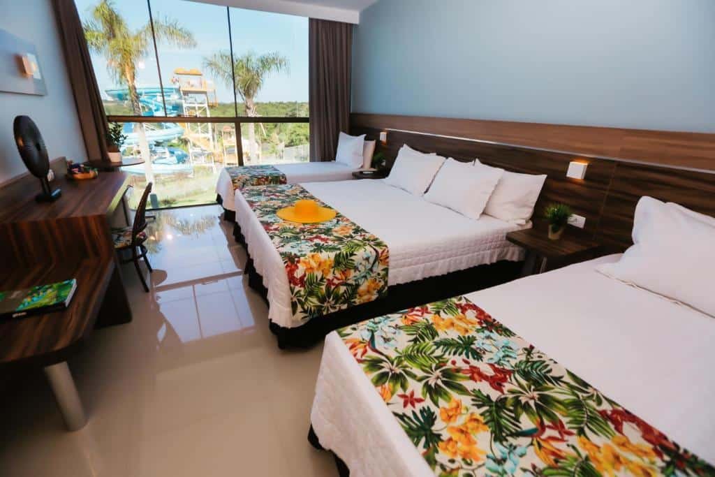 Quarto do Vivaz Cataratas Hotel Resort com três camas lado a lado, sendo elas uma cama de casal e duas camas de solteiro. Em frente a cama de casal tem uma mesa com uma cadeira e do lado da cama de solteiro ao fundo tem uma janela de vidro com vista para a área da piscina.