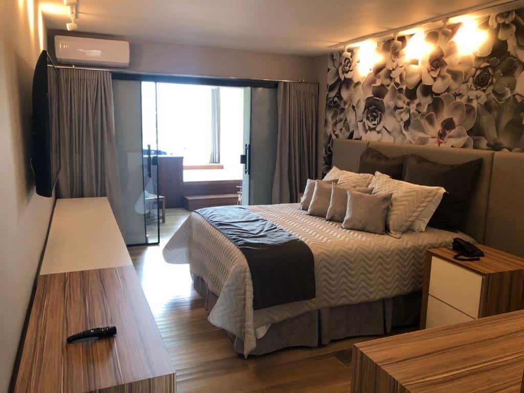 Quarto do Wembley Inn Hotel, uma cama de casal com muitos travesseiros e almofadas, uma sacada ampla, uma ar-condicionado, luzes indiretas sob a cama e uma televisão, para representar pousadas na Praia das Toninhas