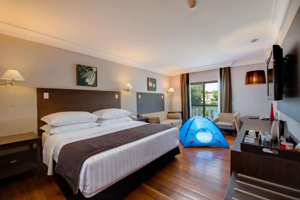 Quarto do Wish Foz do Iguaçu com uma cama de casal e do lado esquerdo um sofá, uma poltrona e uma cabana infaltil. No meio do sofá e da poltrona tem uma porta de vidro com vista para a paisagem. Na frente da cama tem um tv e um armário com objetos em cima.