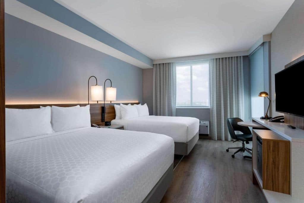 Quarto do Wyndham Garden Miami International Airport com cama de casal do lado direito, uma cômoda com luminária e outra cama de casal do lado esquerdo em frente as camas, cômoda com TV e mesa de Trabalho.