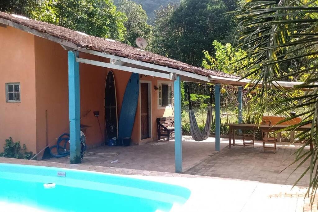 Área externa do Rancho valhalla ubatumirim. Uma piscina no lado esquerdo, no fundo a casa com uma rede, pranchas em pé e uma mesa de madeira com um banco.