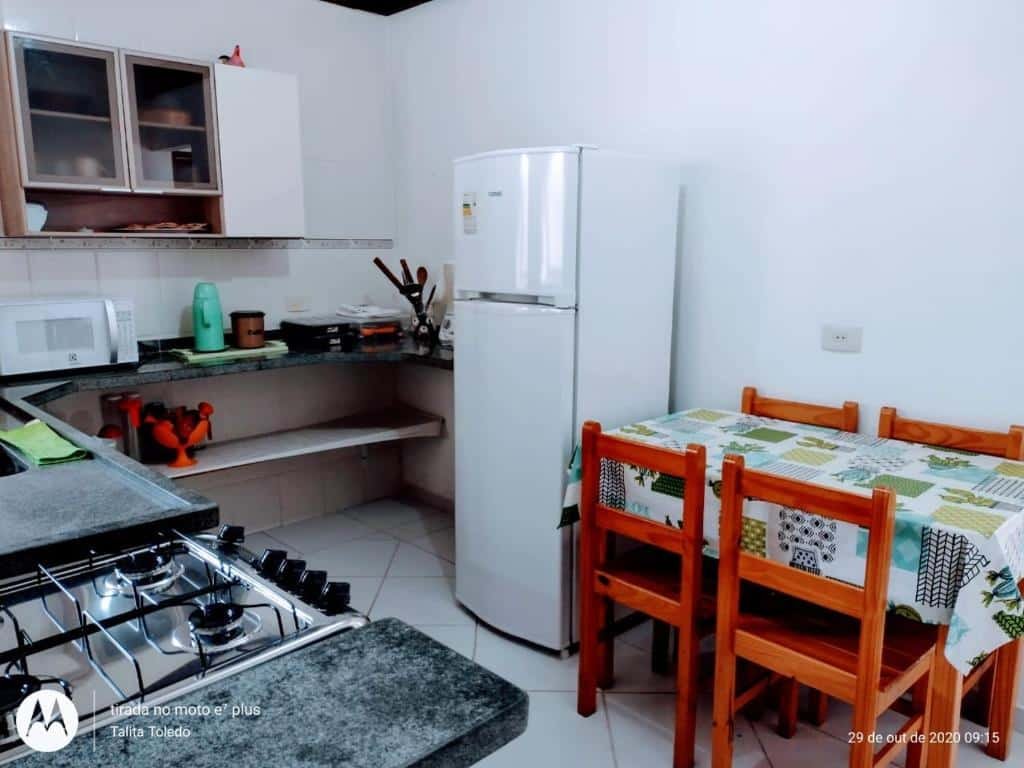 Cozinha do Recanto Canoa Pequena. Uma mesa de madeira com quatro cadeiras do lado direito, do lado uma geladeira. No lado esquerdo na frente a pia e o fogão. No fundo uma bancada com micro-ondas e em cima um armário.